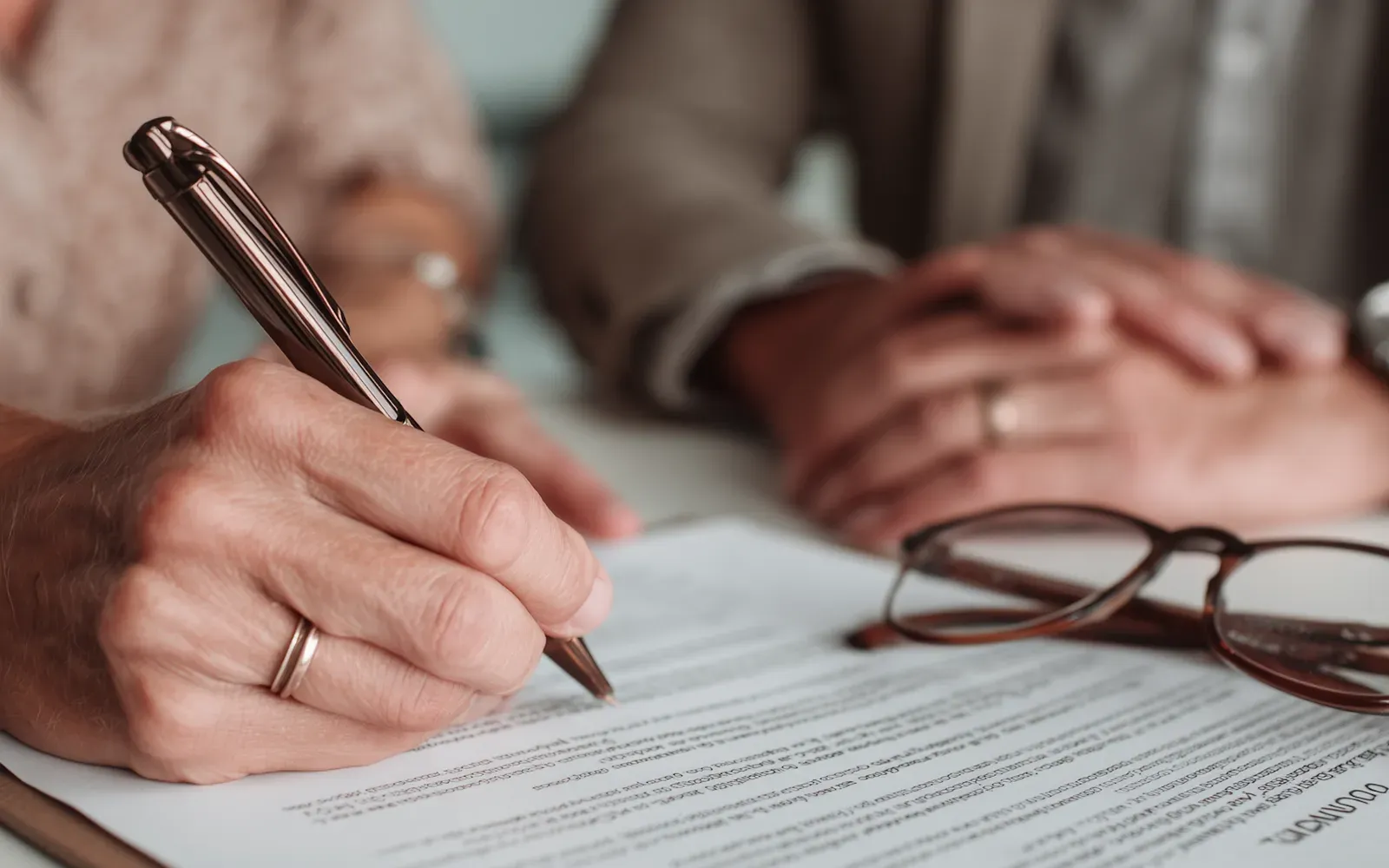 A hand signing a printed agreement with a pen, reading glasses resting on the page.